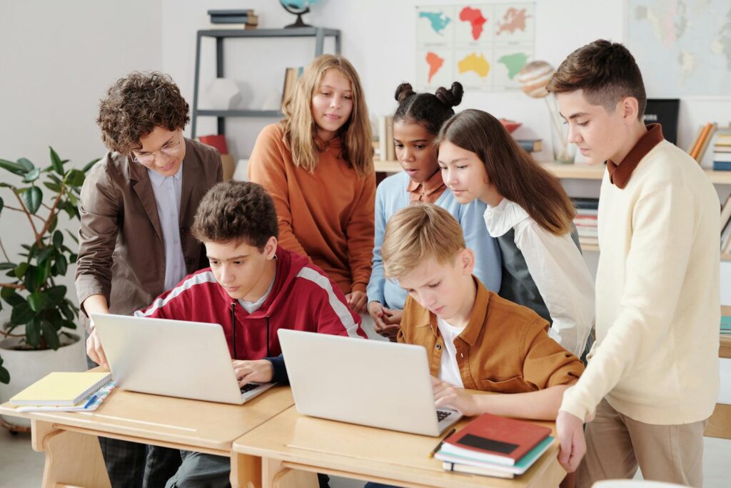 Photo by Max Fischer A group of diverse students collaborating on laptops in a classroom setting, guided by a teacher.